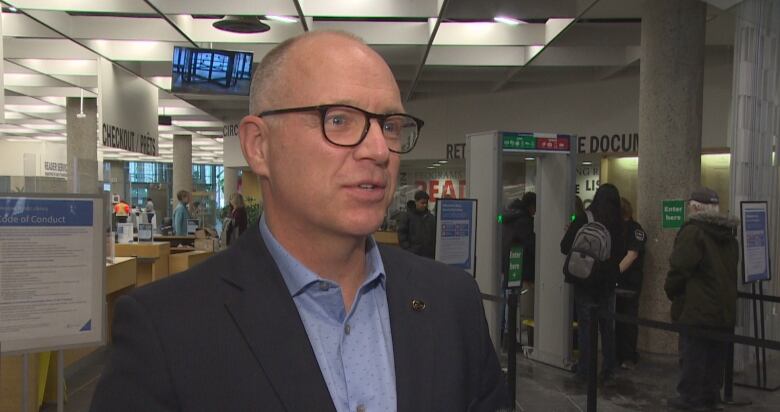 A bald man in glasses, a dark suit jacket and blue collared shirt looks off to the right of the photo frame.