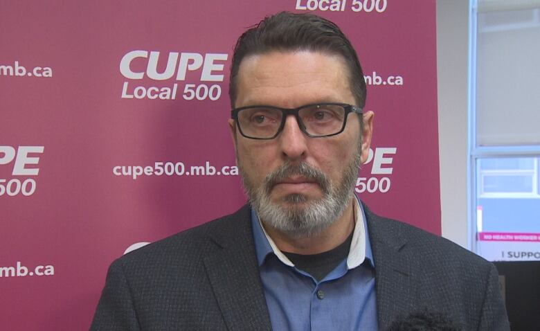 A man in a grey beard and glasses, with a grey suit jacket and blue shirt, is seen from the head and shoulders.