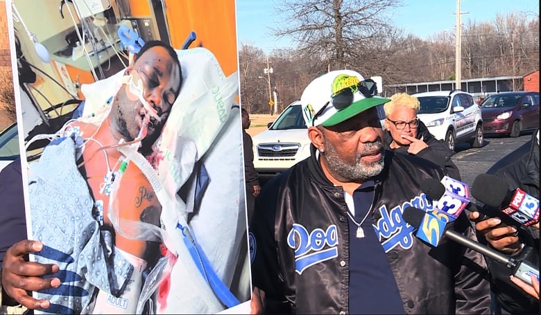 A man wearing a baseball cap and a black jacket speaks into microphones while holding up a large image of a man who is in a hospital bed and connected to tubes.