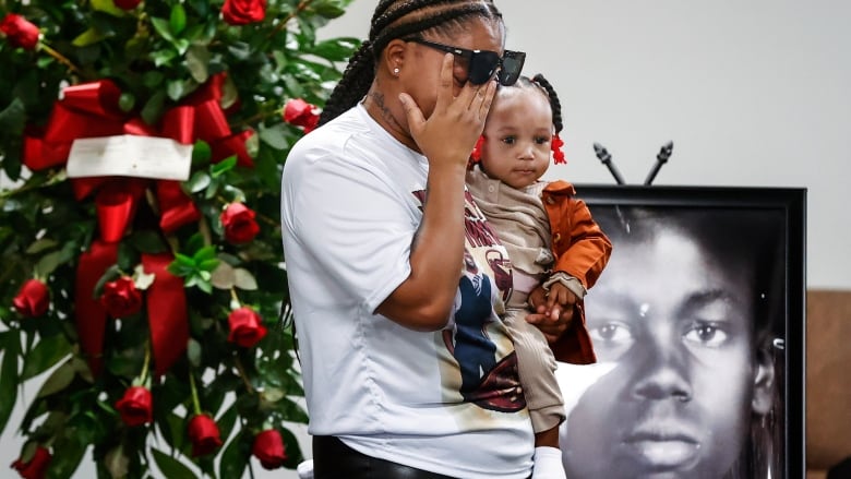 A woman with her hair in braids wearing a white t-shirt and sunglasses wipes her eyes while holding a baby and standing next to a wreath of flowers. Behind her is a black and white image of a man. 