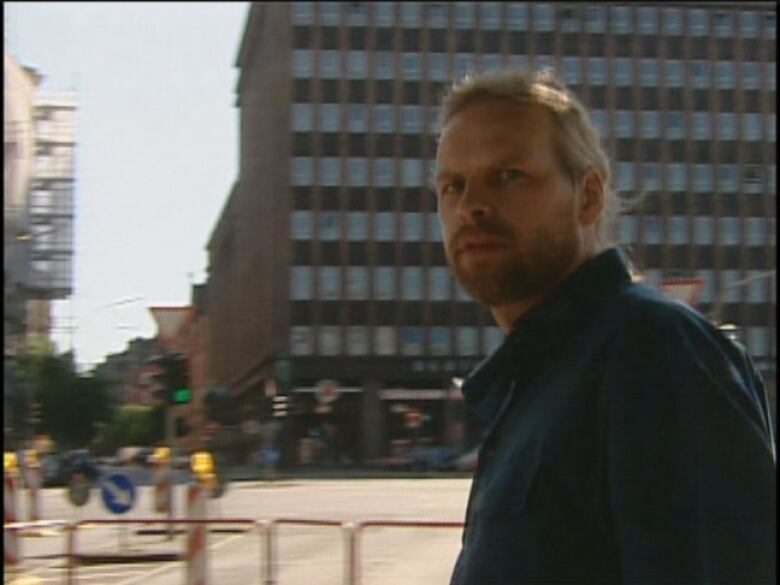 A man on a street turns to look into a camera. 