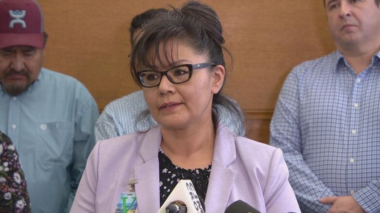 An Indigenous woman is standing. She is wearing glasses, a floral top and a light purple blazer. Her hair is up in a bun. Several men in dress shirts stand behind her. 