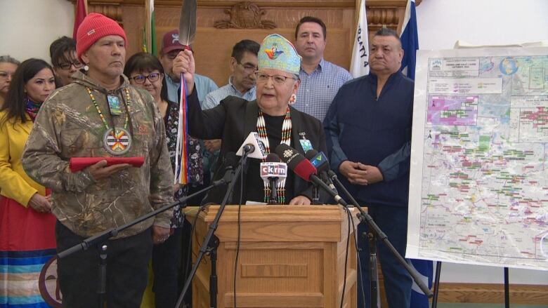 A woman, dressed in black and a traditional headdress, holds up a feather, while speaking into multiple microphones at a podium. To her left is a man, wearing a camouflage hoodie, red toque and ceremonial necklace, who is holding a red cloth. There are many Indigenous women and men standing behind them, in the background. To their right is part of a map of Saskatchewan. 