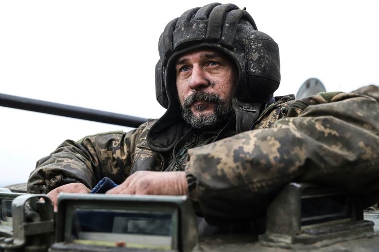 A man with facial wearing a helmet and camoflauge rests his arms at shoulder height on top of the vehicle. 