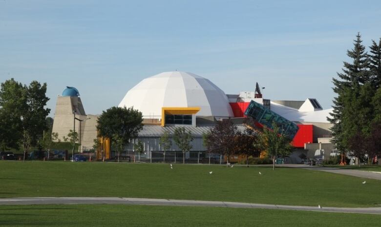 The image shows the angular dome of the Centennial Planetarium in downtown Calgary on a summer day, surrounded by leafy trees and green grass. 