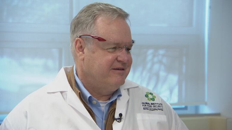 A man wears protective glasses and a white lab coat over his business attire. The logo on the coat reads 'Global Institute for Food Security.'