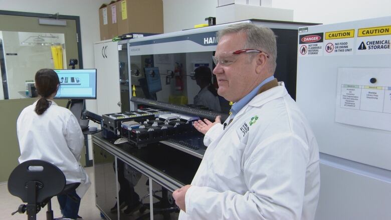 A man wearing protective glasses and a white lab coat motions to a large black machine that has a couple of trays containing small liquid repositories sticking out. Another person in a lab coat works at a computer screen next to the machine.