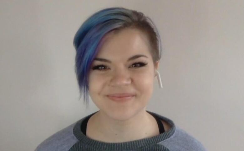 A young person with short blue and purple hair smiles at the camera. 