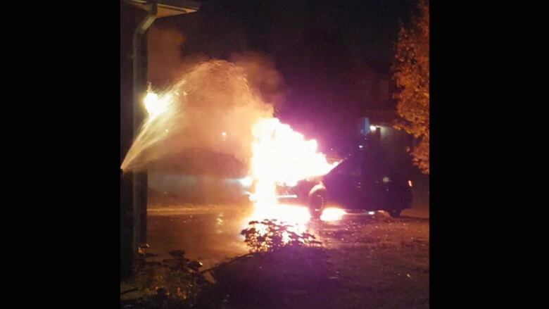 A car parked on a driveway at night is engulfed in flames. A stream of water from a hose is directed at the flames.