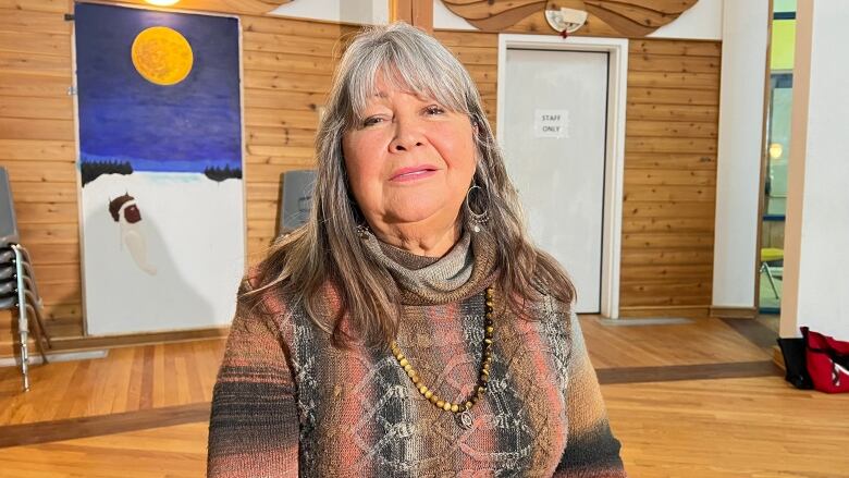 A woman in a brown and pink striped sweater with long grey hair looks at the camera.