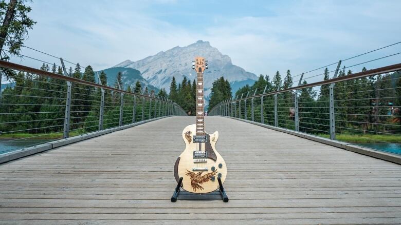 A light-coloured Les Paul-style guitar with the image of an eagle burned into it stands on a bridge over a river before a mountain landscape. 