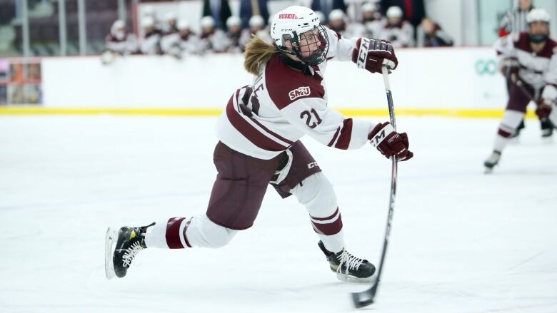 A hockey player with long hair and wearing a dark red and white uniform shoots the puck during a hockey game.