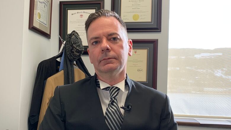 A man wearing a white collared shirt and tie with a black jacket sits at a desk with a view of the ocean from a window in the background. 