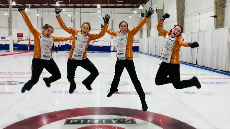 Four women in curling uniforms smile as they jump in the air, with their arms raised, in a curling rink.