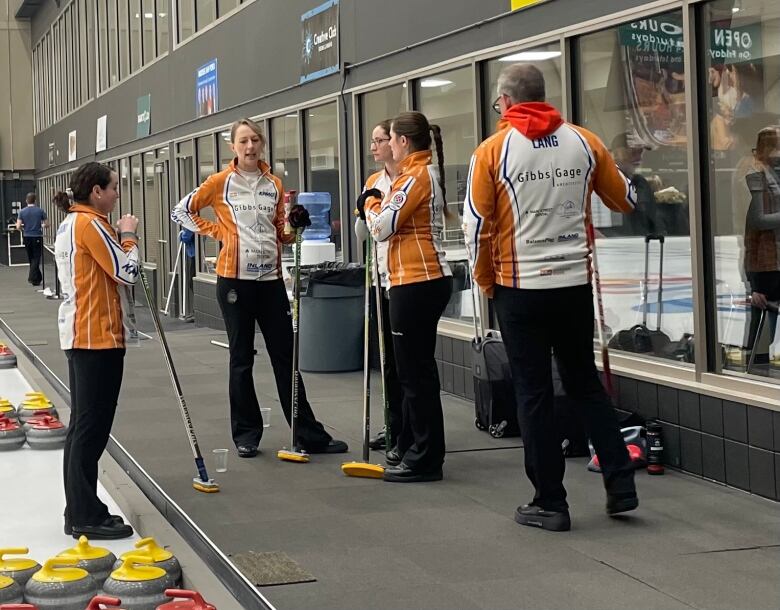 Four women and a man in curling uniforms talk inside a curling rink.