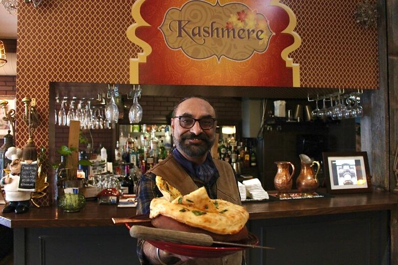 A man holds out a bowl covered in naan to the camera. He stands in front of a restaurant bar with a sign reading 'Kashmere' above him.