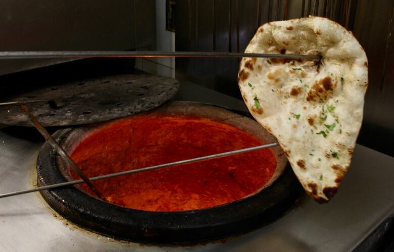 Two metal tongs hold a piece of flatbread over a cylindrical, deep oven glowing deep orange inside.