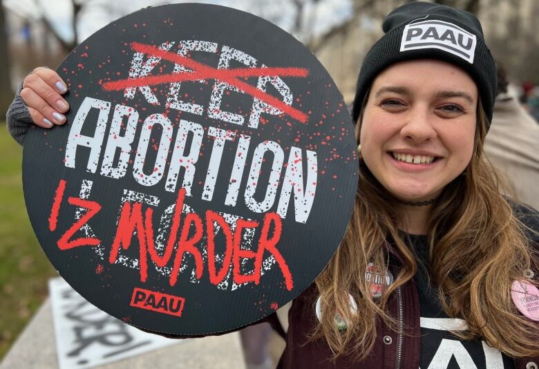 A woman with long hair, wearing a black toque, holds an anti-abortion sign.