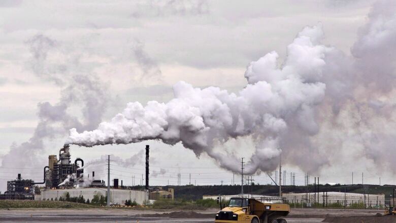 A yellow dump truck drives in front of a mining operation. Smoke from the mine fills the skyline. 