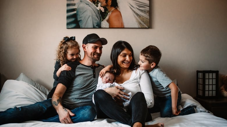 A woman and man sit in bed with their three children. The mother holds her one month old baby in her hand. Behind them, hangs a picture of their wedding day on wall.