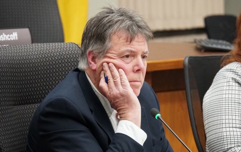 A pensive-looking man wearing a suit sits behind a microphone, his hand resting on his cheek.