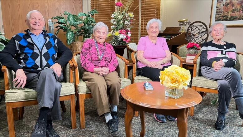 Four elderly people, a man and three women, smile at the camera while sitting in chairs. A small coffee table with a bouquet of flowers stands in the foreground.