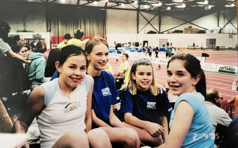 Four adolescent girls smile for a camera. They are sitting in the stands at a track meet, with the track visible behind them.