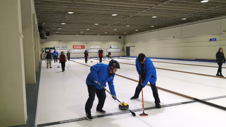 Two men in blue sweep on the ice sheet around a curling stone on an ice sheet.