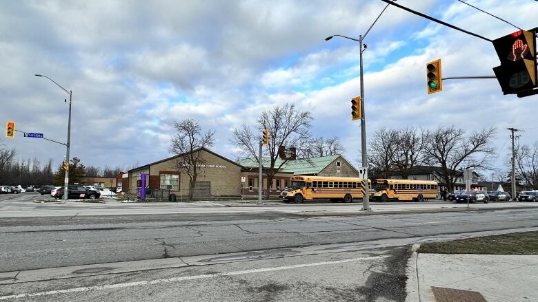 A very wide town street with a school on the far side. school buses and police cars are parked around it.