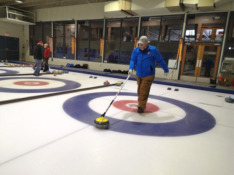 A man in blue jacket uses a broom to organize the stones around the concentric circles on the ice.