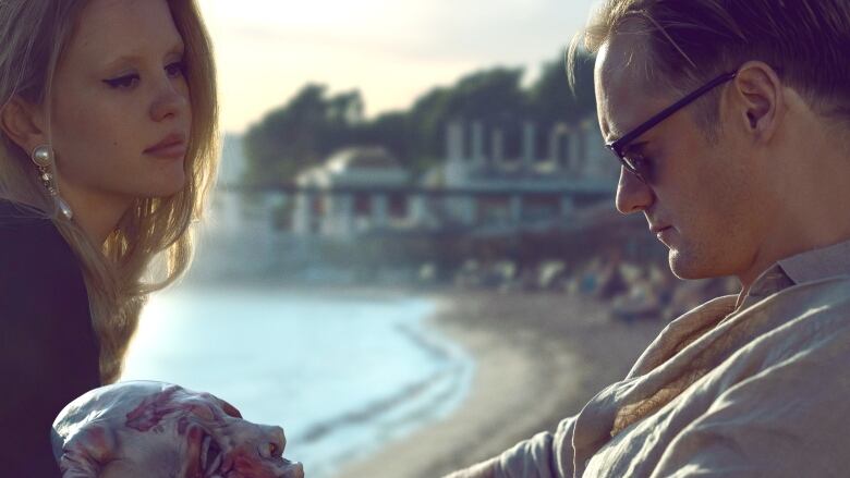 A woman with long blonde hair looks at a man with sunglasses, who stares down at a colourful and disfigured mask. Behind them is a beach. 