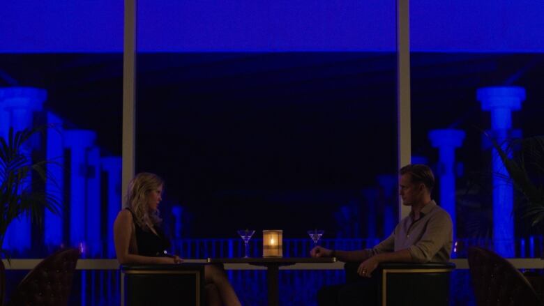 A man and a women sit at opposite sides of a small restaurant table at night. Each have a martini glass in front of them. 