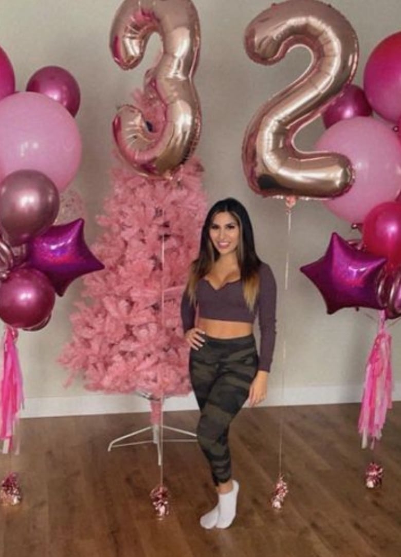 A woman in workout clothes stands with a set of balloons, including two balloons that spell out the number 32.