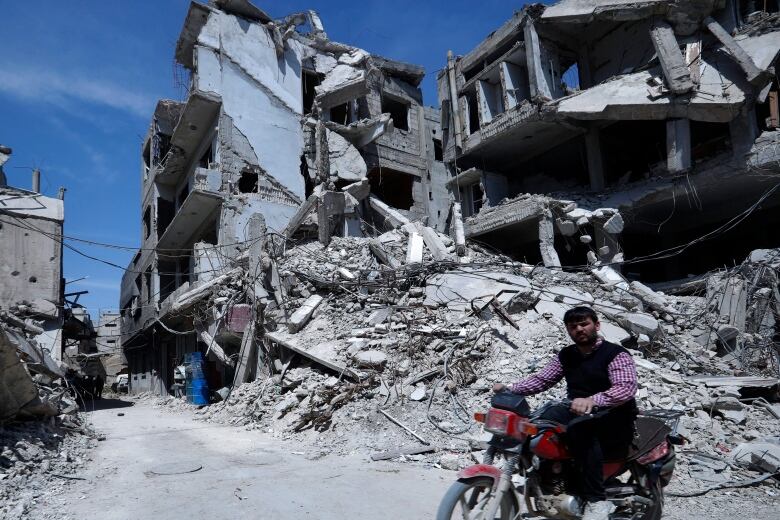 A man on a motorcycle is shown in front of a destroyed building, with rubble on the ground.