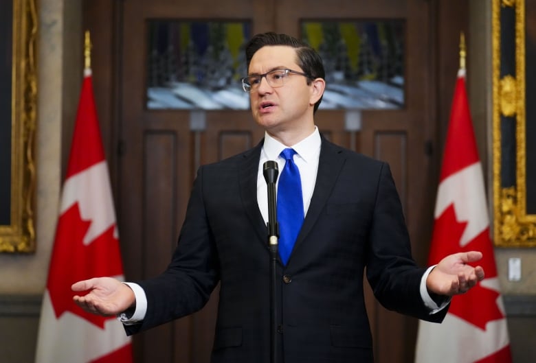 A man in a suit gestures while speaking, flanked on both sides by Canadian flags.