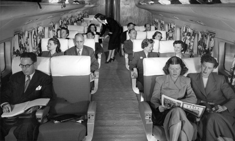 Old black and white photo of men and women sitting in their seats on an airplane. They are dressed formally.