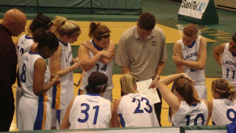 A basketball coach stands in the middle of a team huddle. The players crouch around him as he draws plays on a whiteboard. 