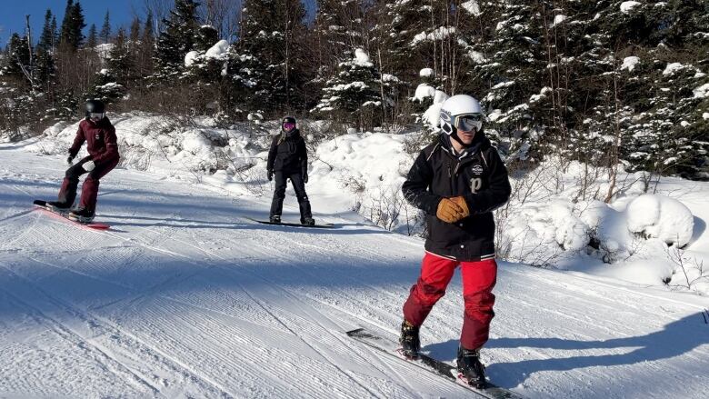 Three snowboarders sliding down a snowy hillside with snow covered trees in the background. 