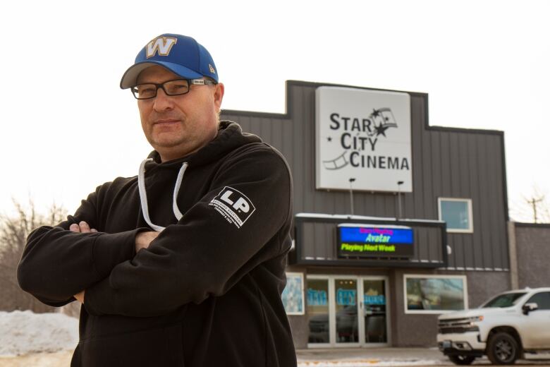 A man wearing a baseball hat stands arms crossed outside a movie theatre.