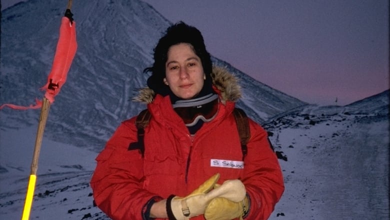 A woman in a red winter parka stands in the snow. Ski goggles rest around her neck. The sky behind her is dark, with a red sunset in the distance.