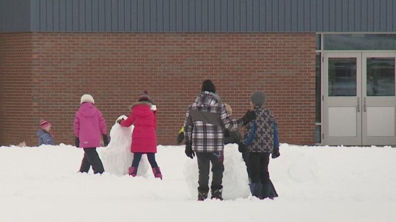 Four children in winter coats building snowmen outside a school building.