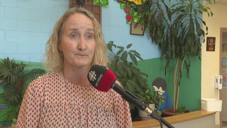 A woman stands in front of a mic, with plants and a mural in the background.