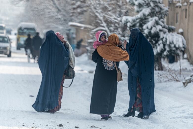 Three women  two of whom are carrying children  stand on a snowy street in Kabul.