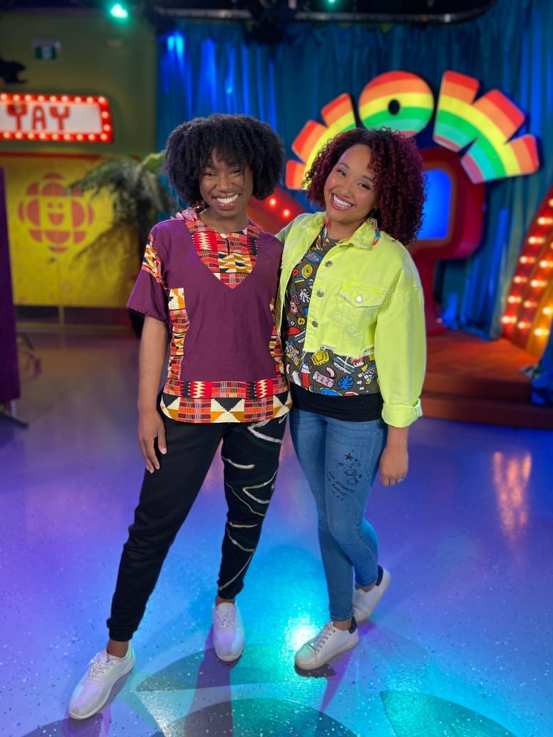 Two Black women with broad smiles, black hair, one wearing a bright yellow jacket and multicoloured shirt the other with a purple shirt trimmed with a pattern of red, orange, black, yellow. Standing in a studio with the word WOW light up in lights in the background.