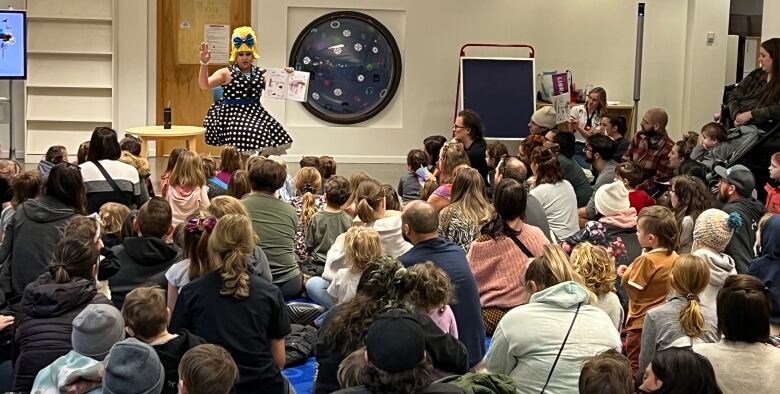 A drag queen wearing a bright yellow wig and polka-dot dress reads to a room full of children and parents.