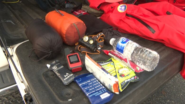 A pick up truck tailgate is loaded with energy bars, a water bottle, flashlight, GPS, sleeping bag, emergency blanket and warm clothing.