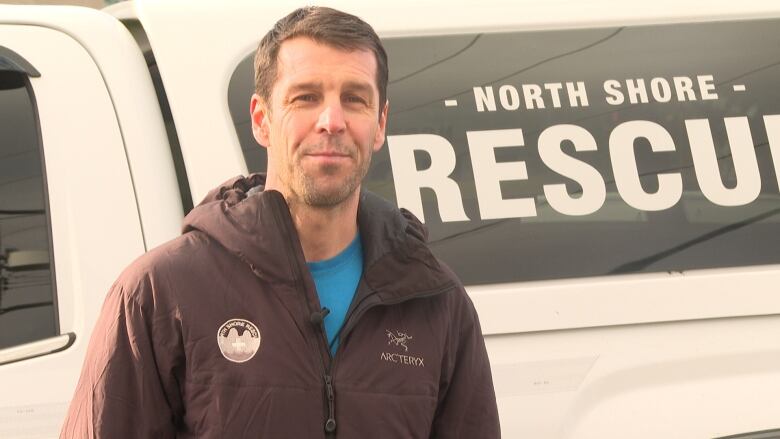 Mike Danks stands in front of a North Shore Rescue truck wearing an Arcteryx jacket. He has short black hair and a chiselled jaw.