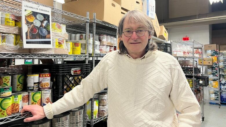 Glenn Rubenstein wearing a white sweater and round glasses standing in front of the food bank's shelves.