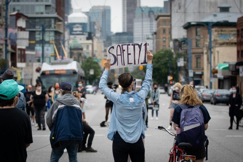 A group of four people stand in the middle of a Vancouver street, with their backs to the camera. One is holding a sign reading 'Safety 1st' above their head.