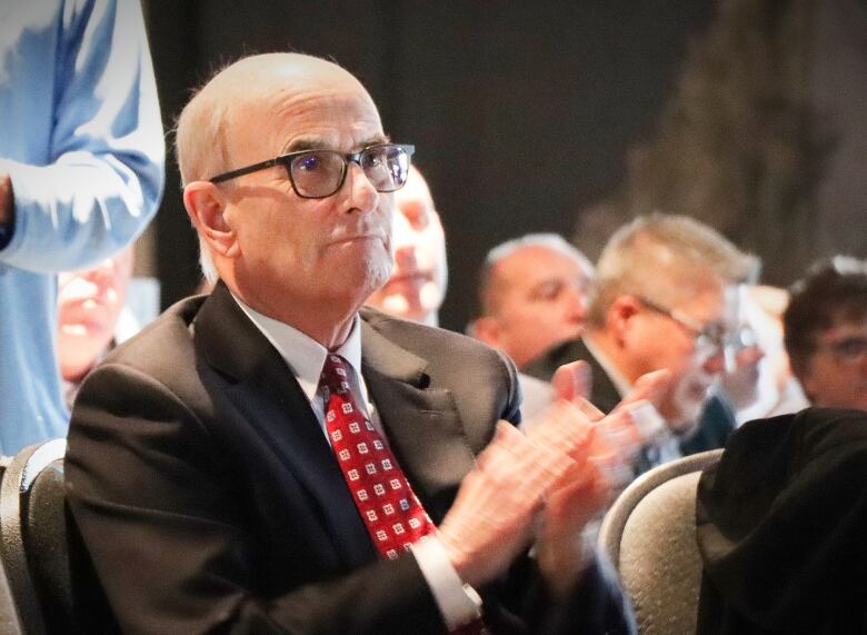 A man in glasses sitting in an audience claps his hands.